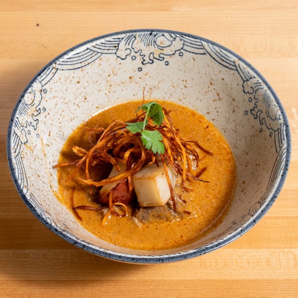 Curry dish with meat, potatoes, fried onions, and cilantro in a decorative bowl on a wooden table.